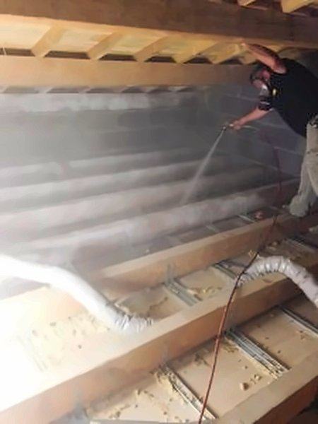 Un technicien traite au pulvérisateur une charpente en bois, dans un nuage de produit.