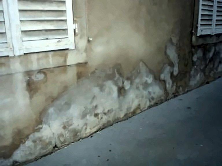 Bas de mur enduit avec du salpêtre et des traces d'humidité qui s'effritent, au pied d'une fenêtre à volets.